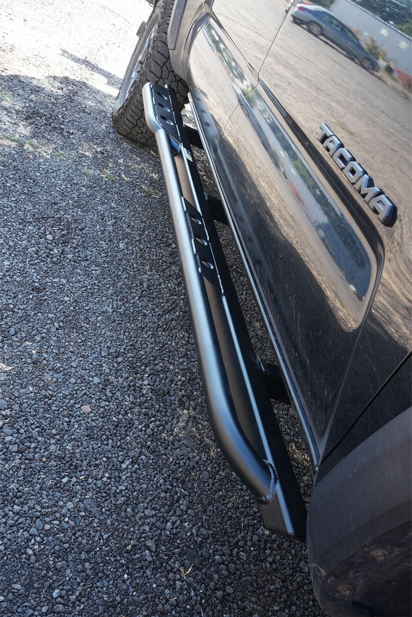Close-up of black metal rock slider installed on 2016 Toyota Tacoma 4-door long bed, showing side step and mounting brackets.