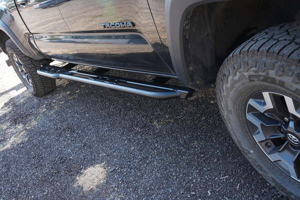 Black metal rock slider installed on the side of a black 2016 Toyota Tacoma 4-door long bed truck.