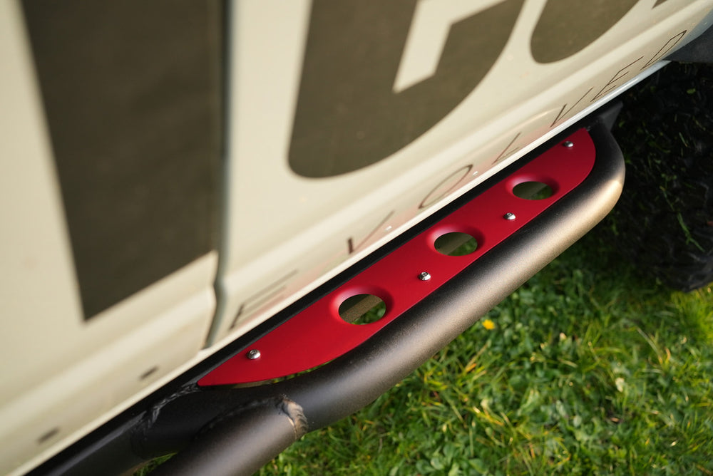 Close-up of red cerakoted step plate mounted on black vehicle side bar with grass background.