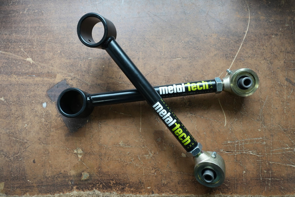 Pair of black Metal Tech rear upper suspension links with metal rod ends, laid crossed on a worn wooden surface.