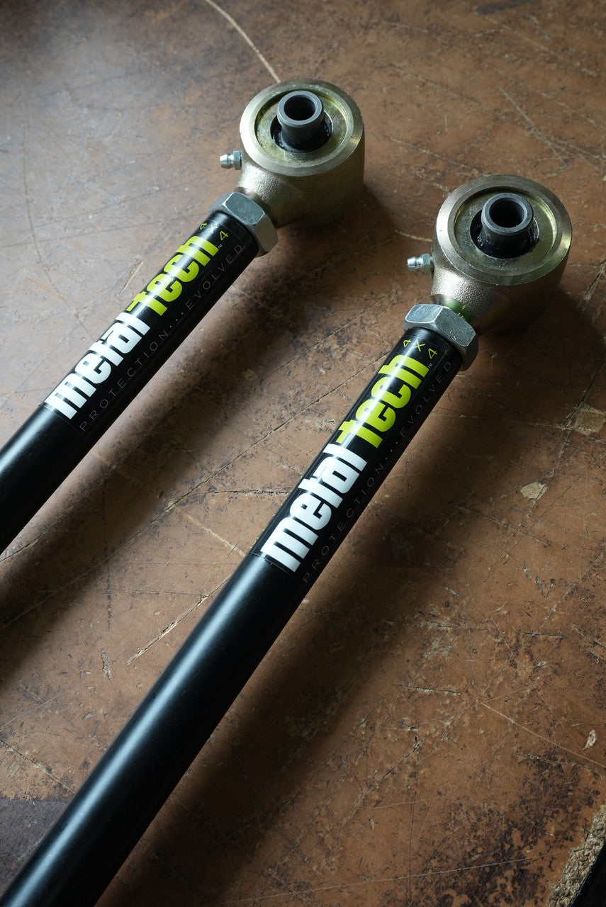 Pair of black Metal Tech rear upper suspension links with metal rod ends, laid crossed on a worn wooden surface.