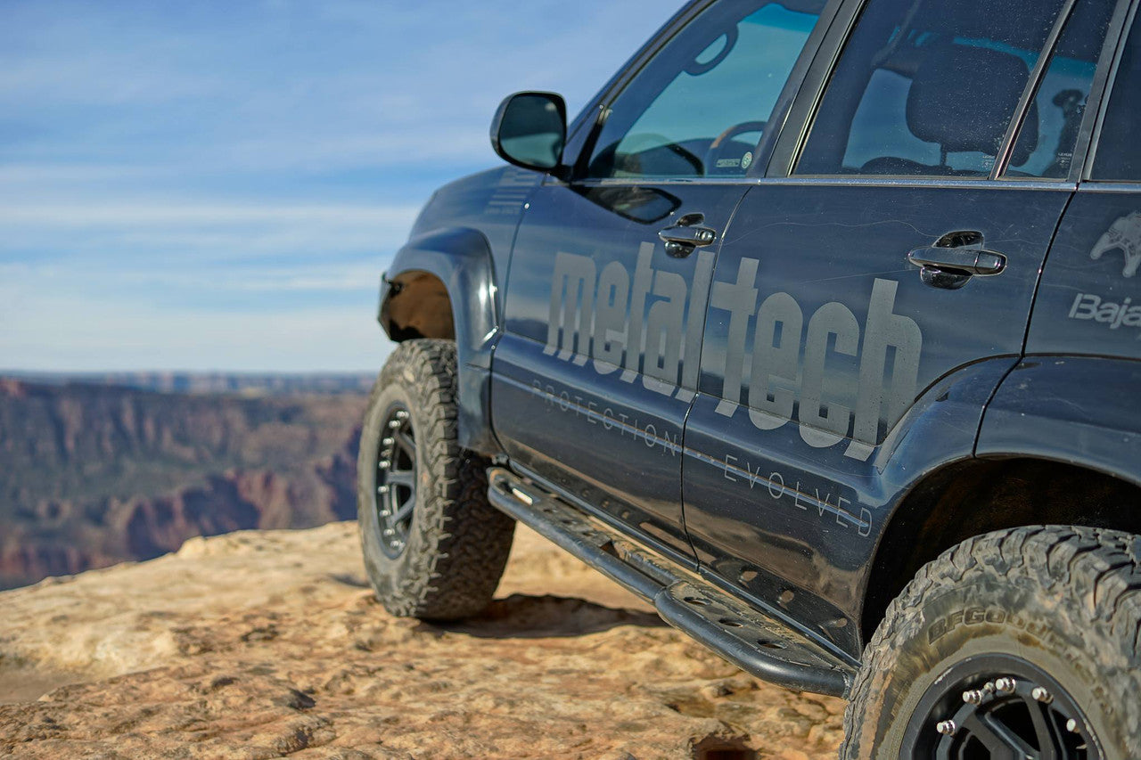 Close-up of black Lexus GX470 with Metal Tech 4x4 rock sliders installed on rocky terrain edge.