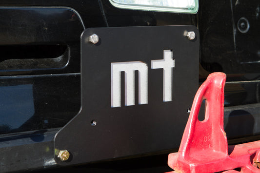 Close-up of black metal license plate holder with cutout "mt" mounted on rear swing-out bumper.