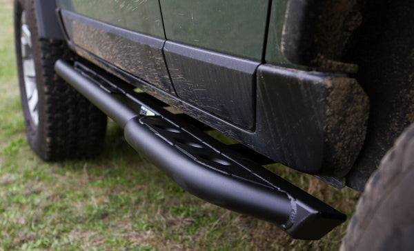 Close-up of black metal rock slider installed on green off-road vehicle side, showing weld details and textured step plate.