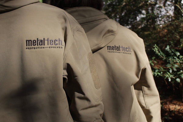 Close-up of two tan Metal Tech 20th Anniversary Tactical Jackets showing embroidered logo on upper back and sleeve patch area.