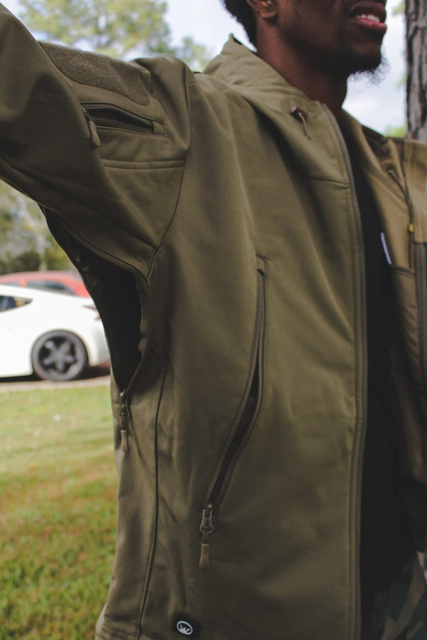 Close-up of olive green tactical jacket sleeve and side zipper pocket on a person outdoors.