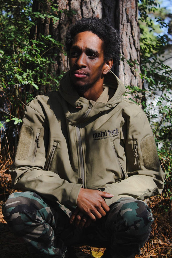 Man wearing olive green Metal Tech 20th Anniversary tactical jacket with hood, sitting outdoors against tree trunk.