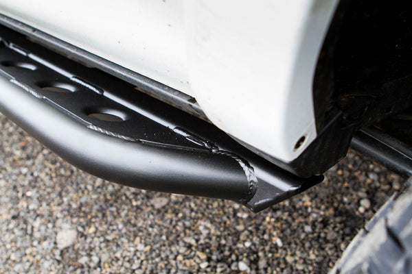 Close-up of black metal trail rock slider installed on the side of a white 2010 4Runner above gravel ground.