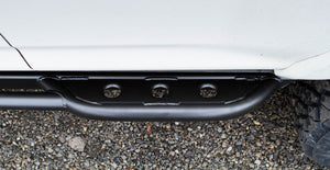 Close-up of black metal rock slider installed on the side of a white 2010 4Runner over gravel ground.