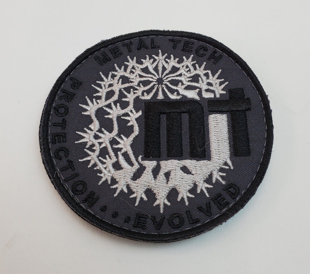 Round black and silver embroidered morale patch with "Metal Tech" text and abstract thorn design.