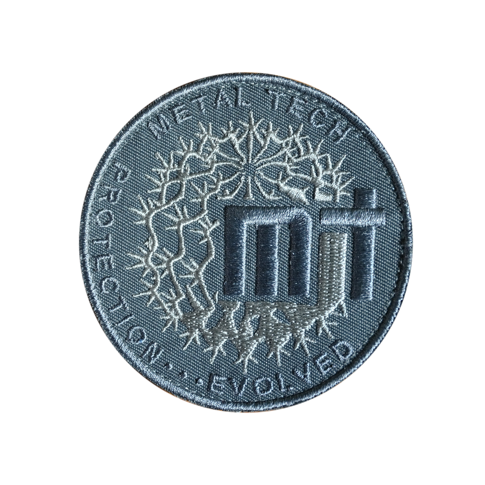 Round embroidered patch with "Metal Tech" text, abstract circuit design, and "Protection...Evolved" slogan in gray tones.