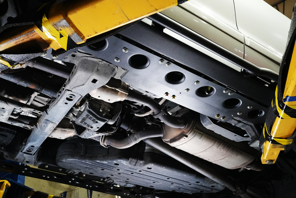 Close-up of metal tech 4x4 rock sliders installed under a white Lexus LX570 showing mounting hardware and exhaust system.