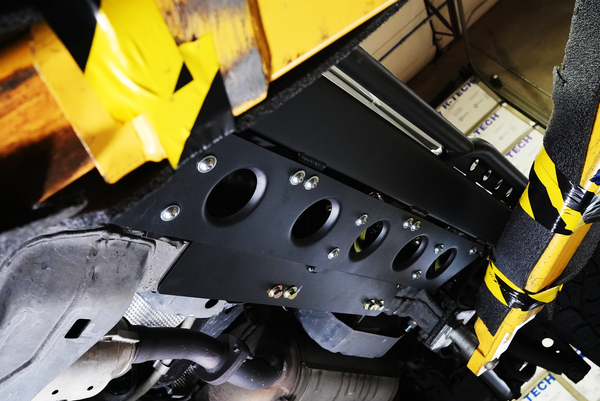 Close-up of black metal rock slider installed under Lexus LX570 chassis, showing mounting bolts and vehicle exhaust system.