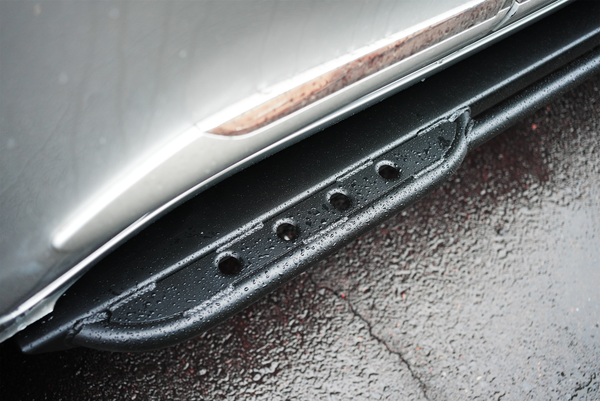 Close-up of black metal rock slider installed on silver Lexus LX570 with water droplets on surface.