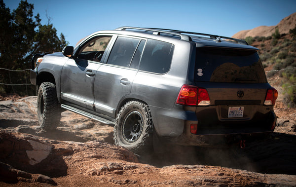 Silver Toyota Land Cruiser with Metal Tech 200 Series rock sliders installed, driving over rocky terrain outdoors.