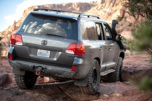 Rear side view of gray 2008-2021 Toyota Land Cruiser with Metal Tech 200 Series rock sliders on rocky terrain.
