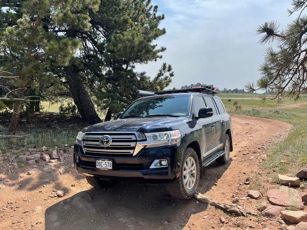 Black 200 Series Toyota Land Cruiser with Metal Tech rock sliders installed, parked on a dirt trail in a forested area.