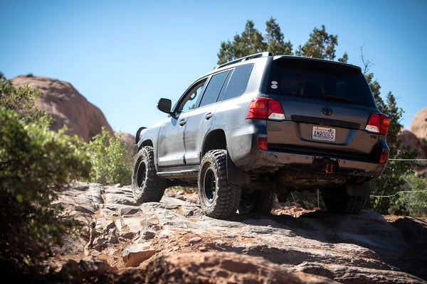 Rear side view of a Toyota Land Cruiser with Metal Tech 200 Series rock sliders driving on rocky terrain.