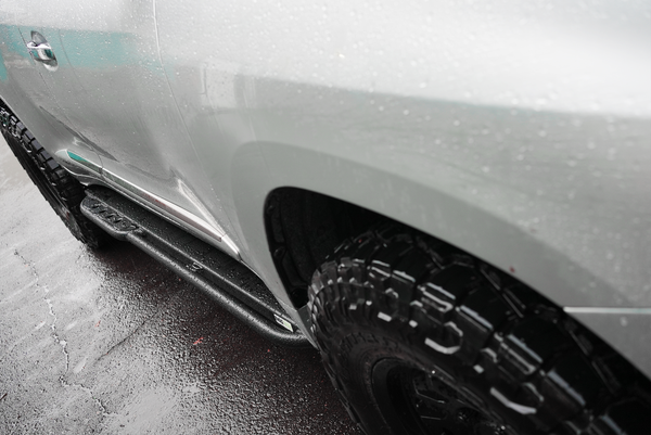 Close-up of black Metal Tech 4x4 rock slider installed on silver Lexus LX570 with wet pavement background.