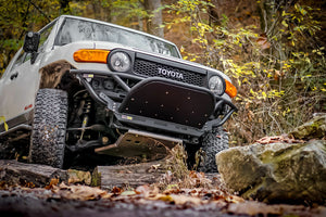 White Toyota FJ Cruiser with Metal Tech front tube bumper driving over rocky trail in forest setting.