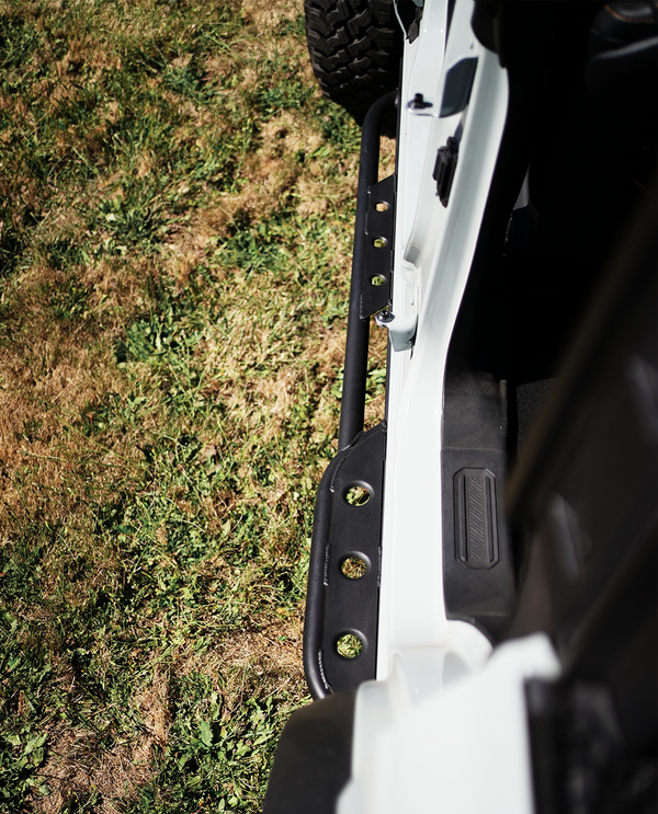 Close-up of black metal rock slider installed on the side of a white 2021 Ford Bronco 4-door on grass.