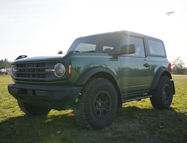2021 green Ford Bronco 2-door with Sidewinder RRR rock sliders installed, parked on grass in sunlight.
