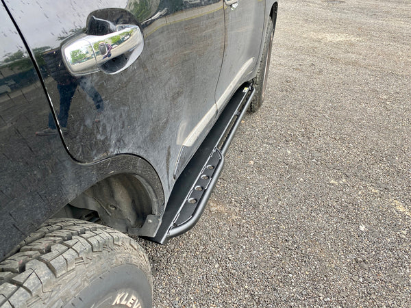 Close-up of black metal rock slider installed on the side of a black Lexus LX570 with off-road tires.