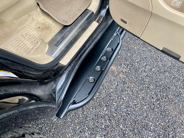 Close-up of black metal rock slider installed on the side of a Lexus LX570 with open door and gravel ground.