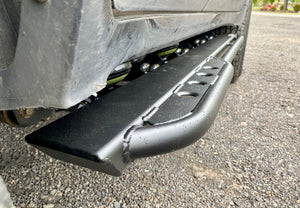 Close-up of black metal rock slider installed on Lexus LX570, showing welds and textured finish on gravel surface.