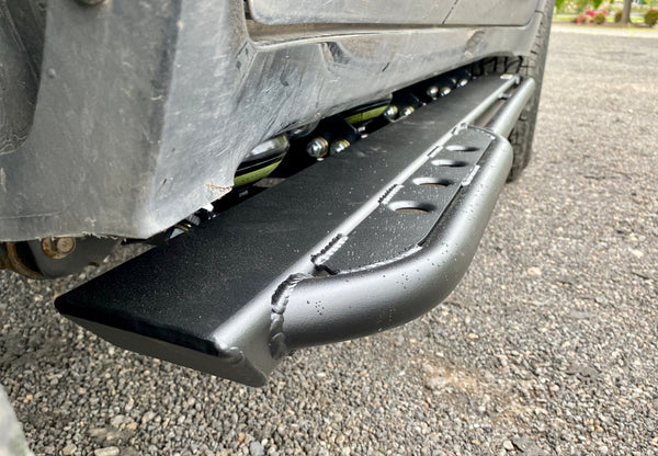 Close-up of black metal rock slider installed on Lexus LX570, showing welds and textured finish on gravel surface.