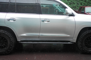 Side view of silver Lexus LX570 with black Metal Tech 4x4 rock sliders installed on wet pavement.