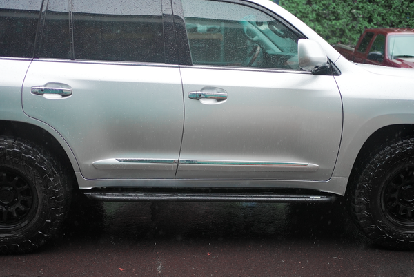 Side view of silver Lexus LX570 with black Metal Tech 4x4 rock sliders installed on wet pavement.