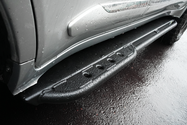 Close-up of black Metal Tech 4x4 rock slider installed on silver Lexus LX570, wet from rain, showing textured surface and mounting.