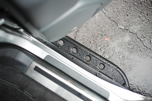 Close-up of black metal rock slider with drainage holes installed on Lexus LX570 door sill with water droplets.