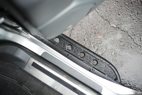 Close-up of black metal rock slider with drainage holes installed on Lexus LX570 door sill with water droplets.