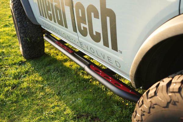 Close-up of black and red Sidewinder rock sliders installed on the side of a 2021 Bronco 4-door on grass.