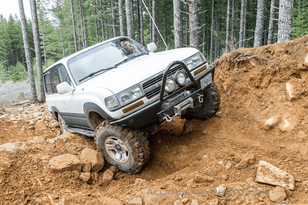 White 80 Series Land Cruiser with Metal Tech 4x4 rock sliders climbing rocky off-road terrain in forest.