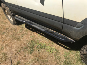 Black metal rock slider installed on the side of a white 80 Series Land Cruiser on dry grass.