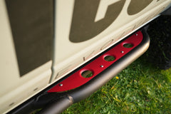 Close-up of red cerakoted step plate mounted on black vehicle side bar with grass background.