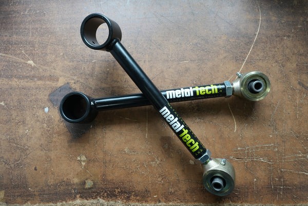 Pair of black Metal Tech rear upper suspension links with metal rod ends, laid crossed on a worn wooden surface.