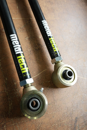 Close-up of two black Metal Tech rear upper suspension links with metal rod ends on a brown surface.