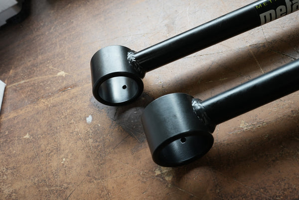 Close-up of two black Metal Tech rear upper links with visible welds and mounting holes on a worn surface.