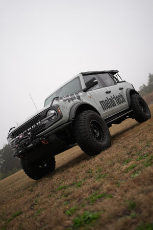 2021 Ford Bronco 4-door with Metal Tech rock sliders installed, shown from front side on grassy terrain.