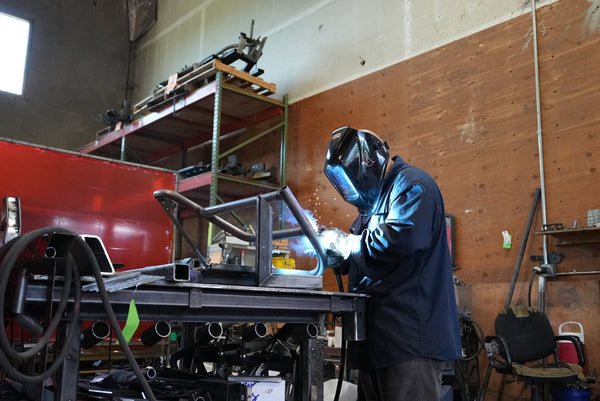 Welder fabricates metal tube door frame for FJ45 Land Cruiser in workshop setting.