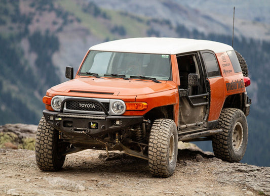 Orange Toyota FJ Cruiser with Metal Tech Phantom front bumper, off-road tires, and open driver door on rocky terrain.