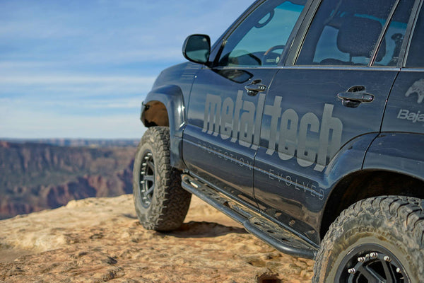 Close-up of black Lexus GX470 with Metal Tech 4x4 rock sliders installed on rocky terrain edge.