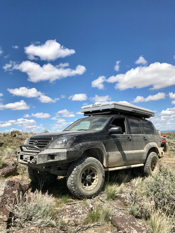 Black Lexus GX470 with roof tent and metal rock sliders driving off-road on rocky terrain under blue sky.