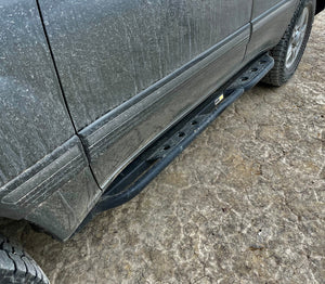 Black metal rock slider installed on the side of a dirty 100 Series Land Cruiser LX470 on cracked dirt ground.