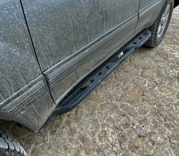 Black metal rock slider installed on the side of a dirty 100 Series Land Cruiser LX470 on cracked dirt ground.