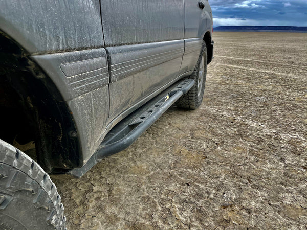 Close-up of black rock slider installed on dirty 100 Series Land Cruiser LX470 side in dry cracked terrain.